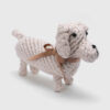 Brinquedo de corda em formato de cachorro, cor clara, com laço marrom e detalhes bordados, sobre fundo branco